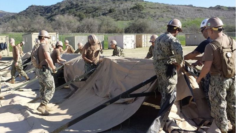 Soldiers in desert camouflage uniforms erecting a large beige tent at an outdoor base, with containers and hills in the background.